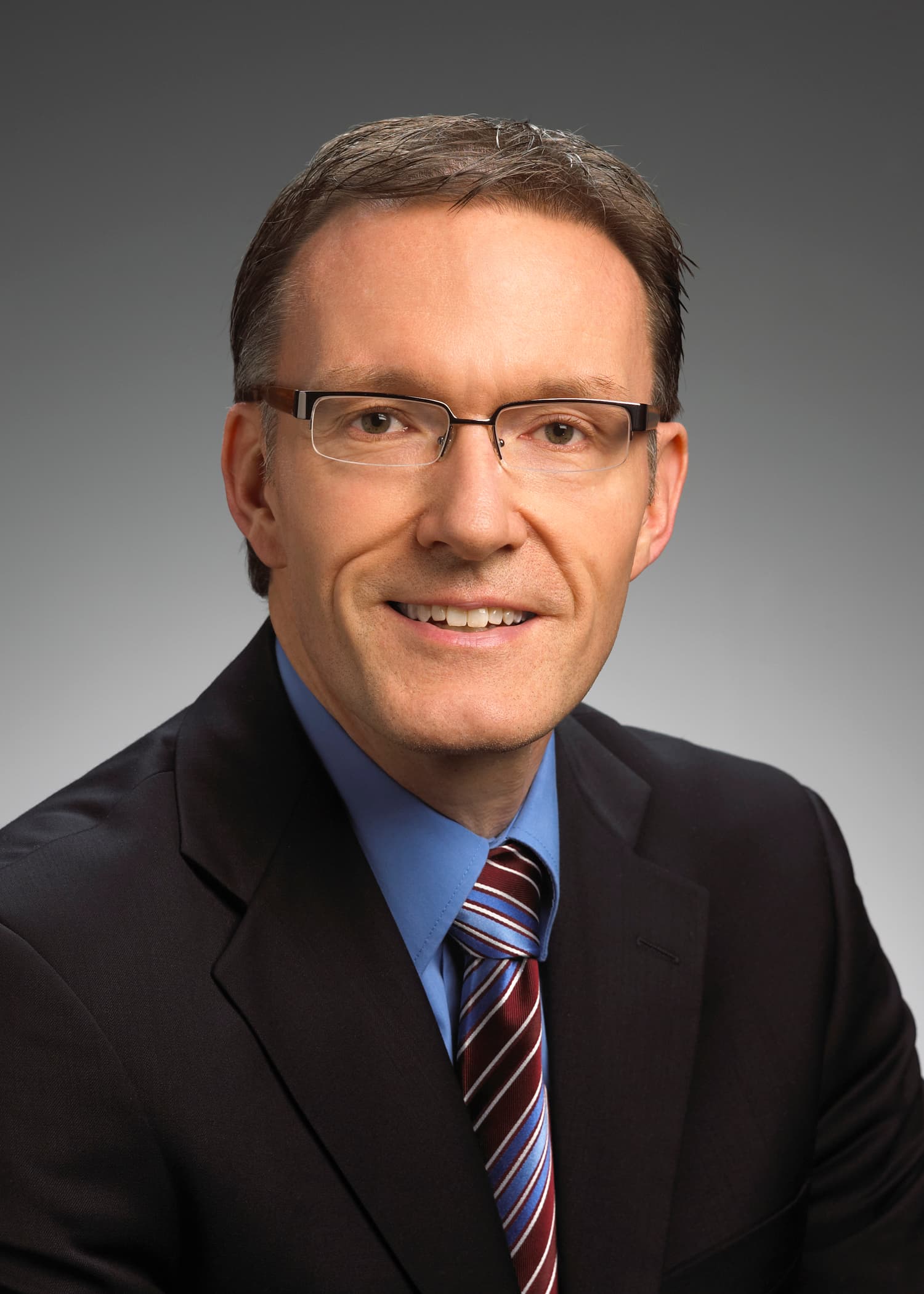 Man in a suit and tie with glasses, smiling, in front of a plain gray studio background.